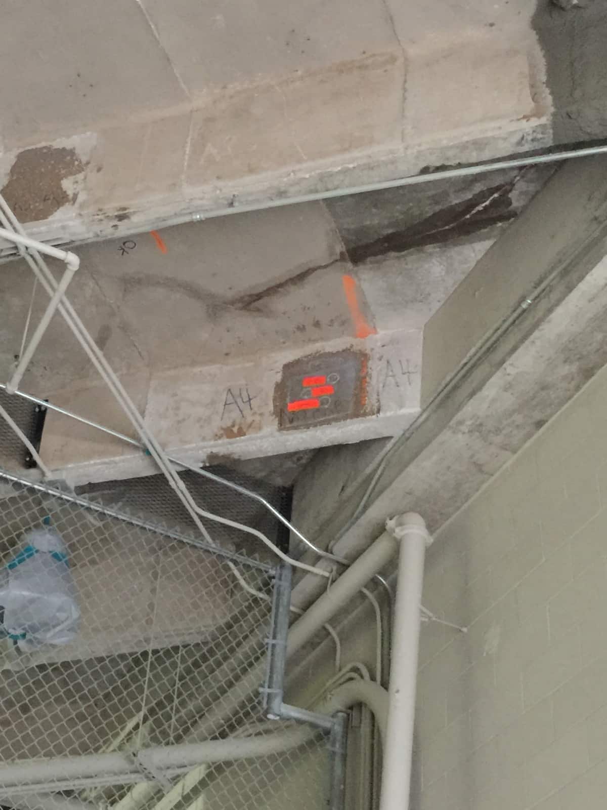 Concrete beams with orange paint markings and a patch above pipes and chain-link.