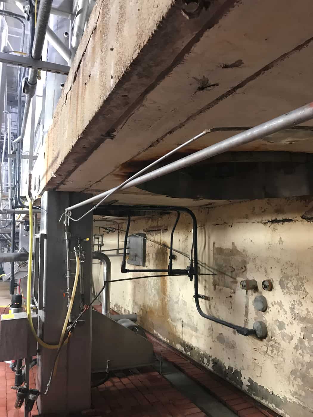 Deteriorated concrete beam with rust staining above a brick-tile floor and piping.