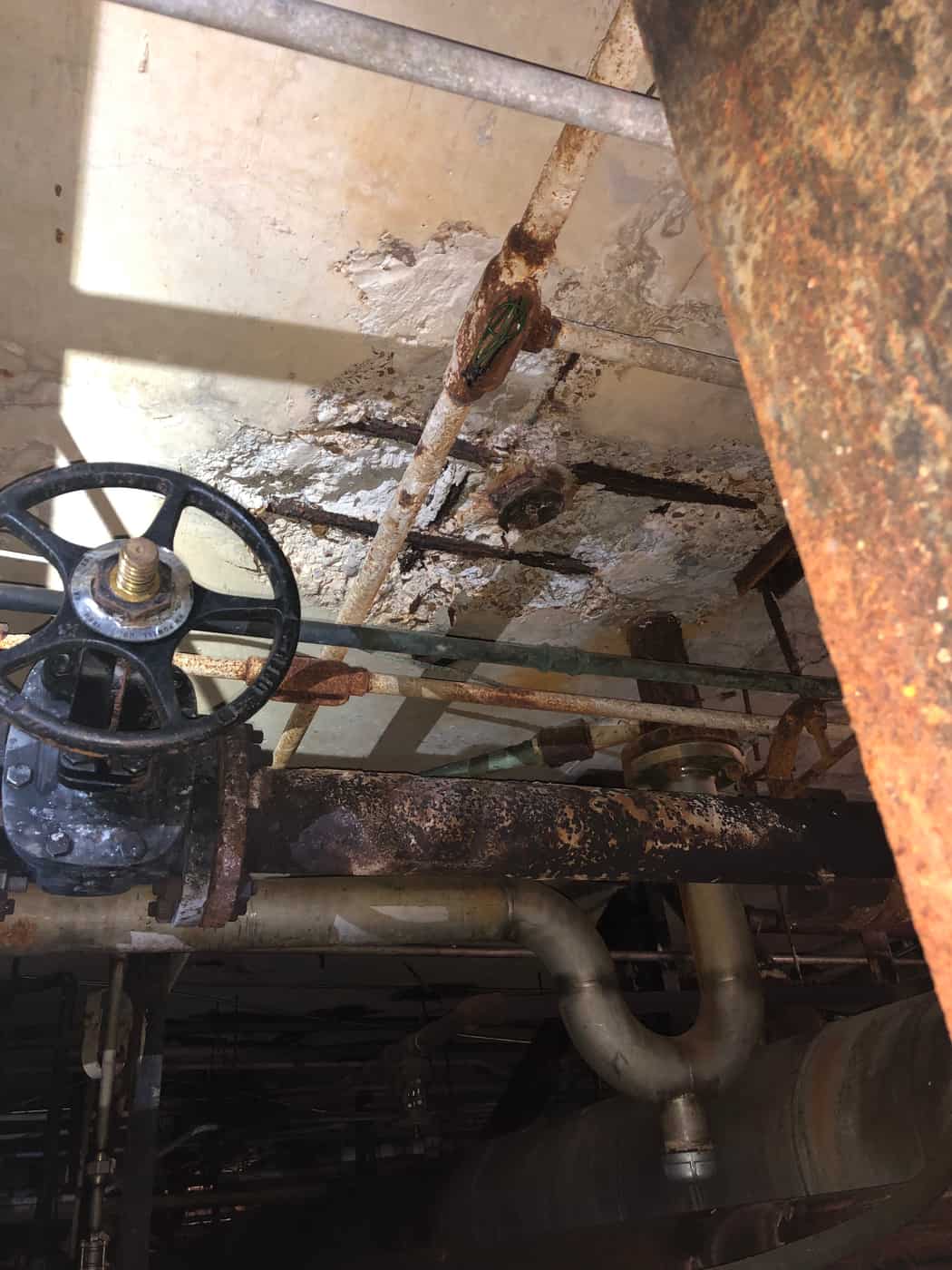 Spalled slab and beam with rusted rebar above a gate valve and piping.