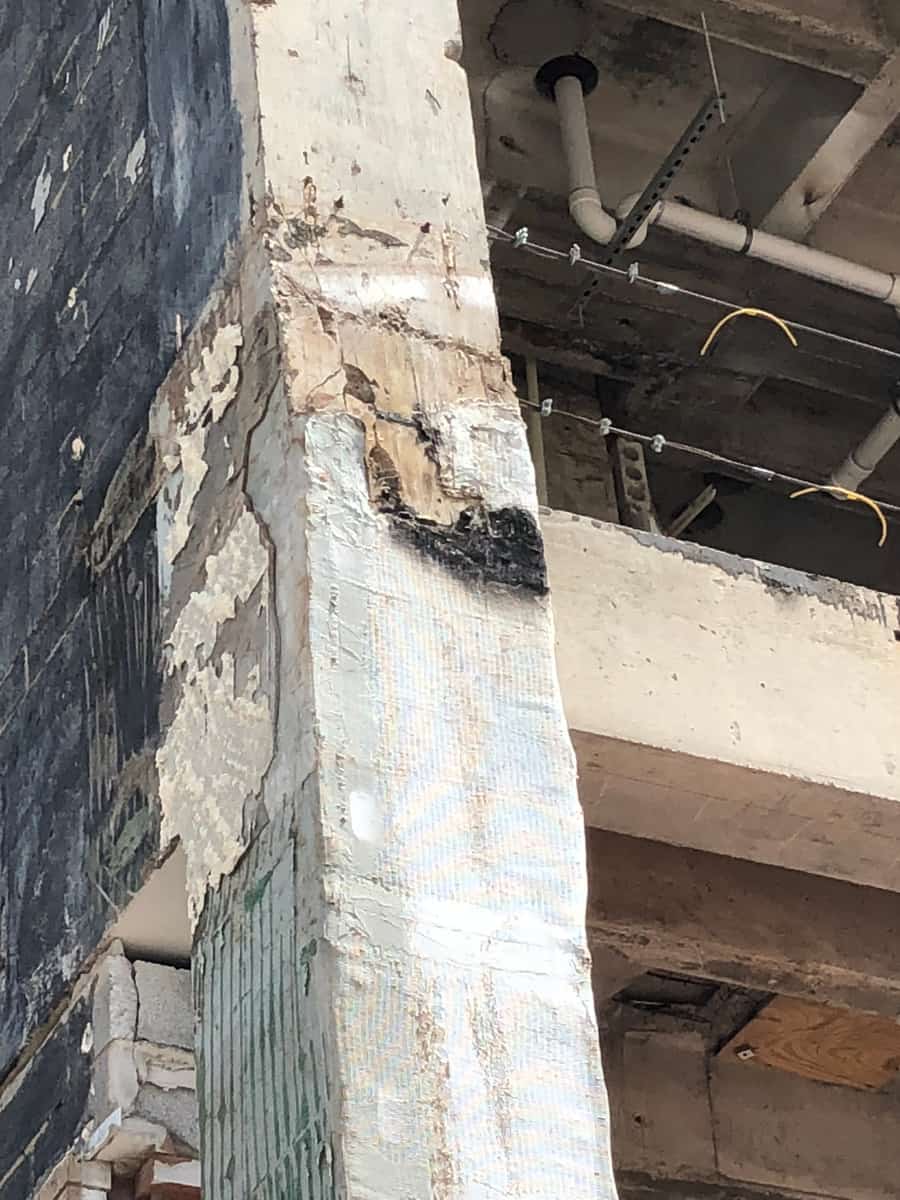Deteriorated concrete column corner with spalled, peeling surface beside a brick wall.