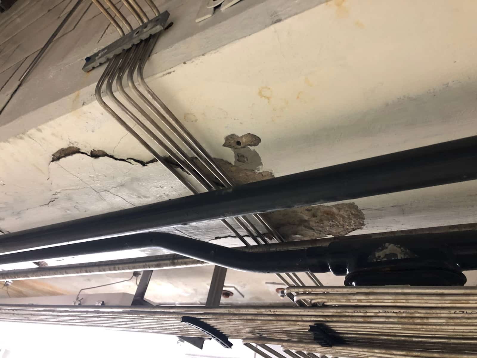 Concrete beam with a crack and a spalled patch behind conduits.