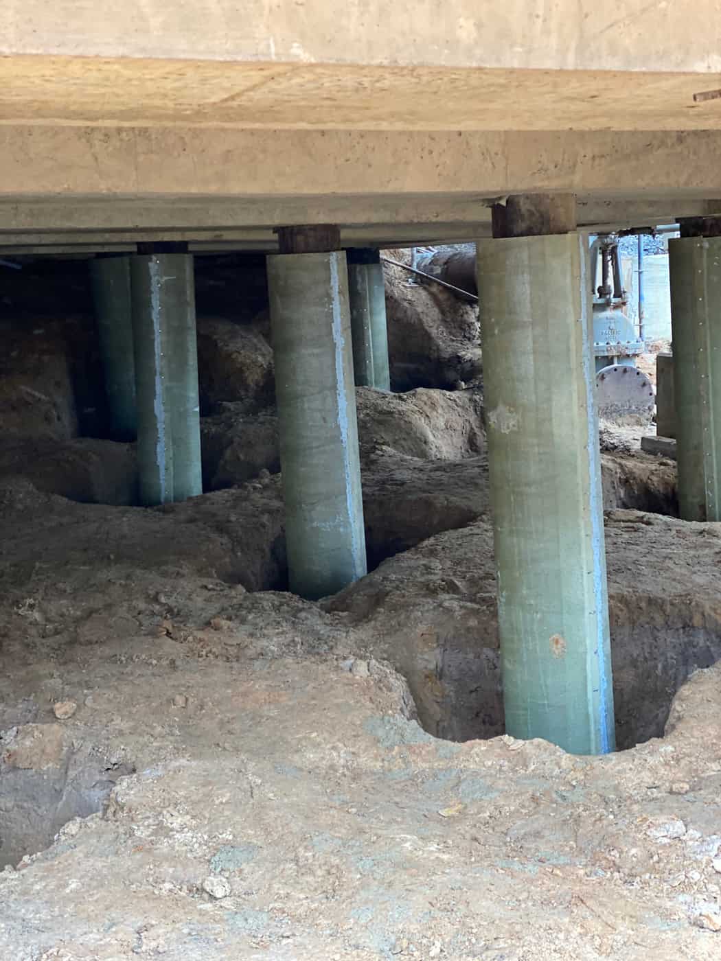 Several fiberglass-jacketed piles set in excavated holes beneath a concrete structure.