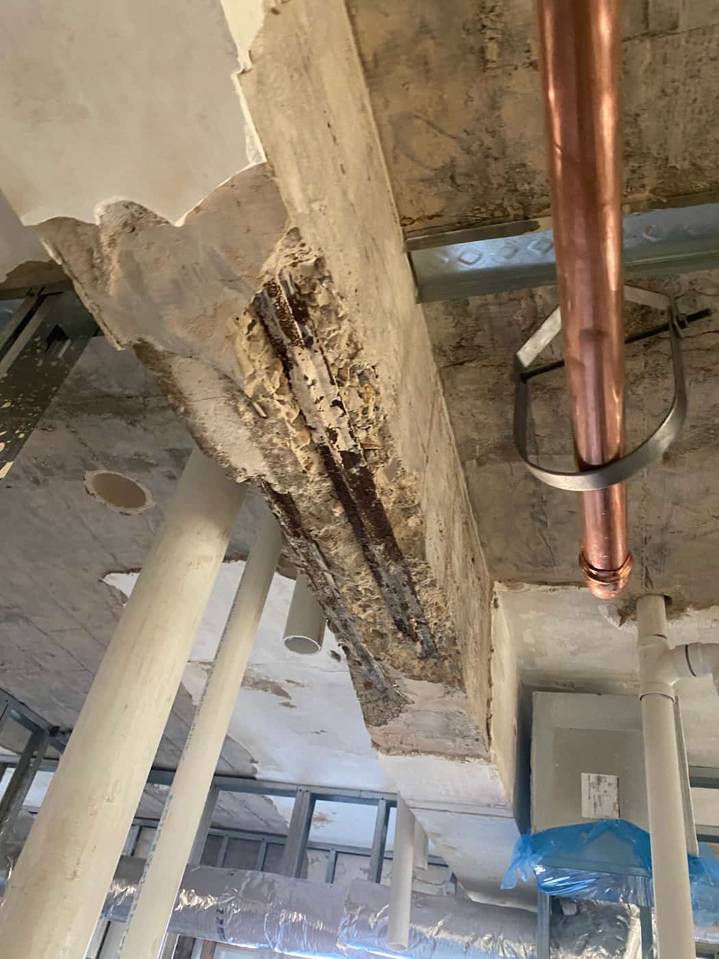 Deteriorated beam with exposed corroded rebar next to a copper pipe.