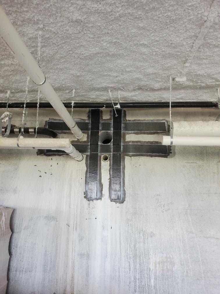 Black composite strips framing a penetration below pipes on a concrete surface.
